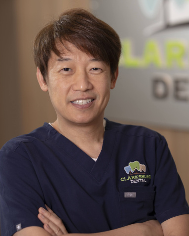 The image shows a man wearing a white lab coat, standing in front of a sign with the word Clear on it, posing for a professional headshot.