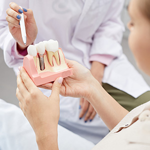 The image shows a dentist holding up a model mouth with teeth and implants for demonstration purposes, while another person observes closely.